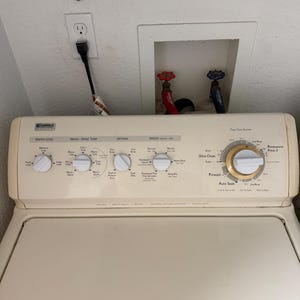 May include: A cream-colored washing machine with a control panel featuring knobs for water level, wash/rinse temperature, and options. The panel includes settings for speeds and a dial for prewash and auto soak. Above the machine are water valves and an electrical outlet.