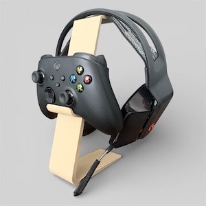 Könnte beinhalten: Beigefarbener Holzständer für Gaming-Headset und Controller. Der Ständer hält einen schwarzen Xbox-Controller mit farbigen Knöpfen und ein schwarzes Gaming-Headset. Das Headset hat ein schwarz-graues Kopfband und ein schwarzes Mikrofon.
