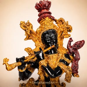 Puede incluir: Una estatua negra y dorada de una figura tocando la flauta, con una corona roja y un pavo real rojo posado en el hombro. La figura tiene joyas y tocados dorados ornamentados. El texto DivijaSacredArt está impreso en la base de la estatua.