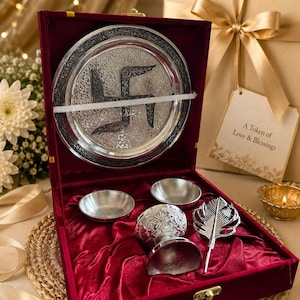May include: A silver-toned gift set in a red velvet-lined box containing a circular plate with an engraved swastika, two small bowls, a pedestal cup, and a feather-shaped spoon. A gift tag reads A Token of Love & Blessings in black text.