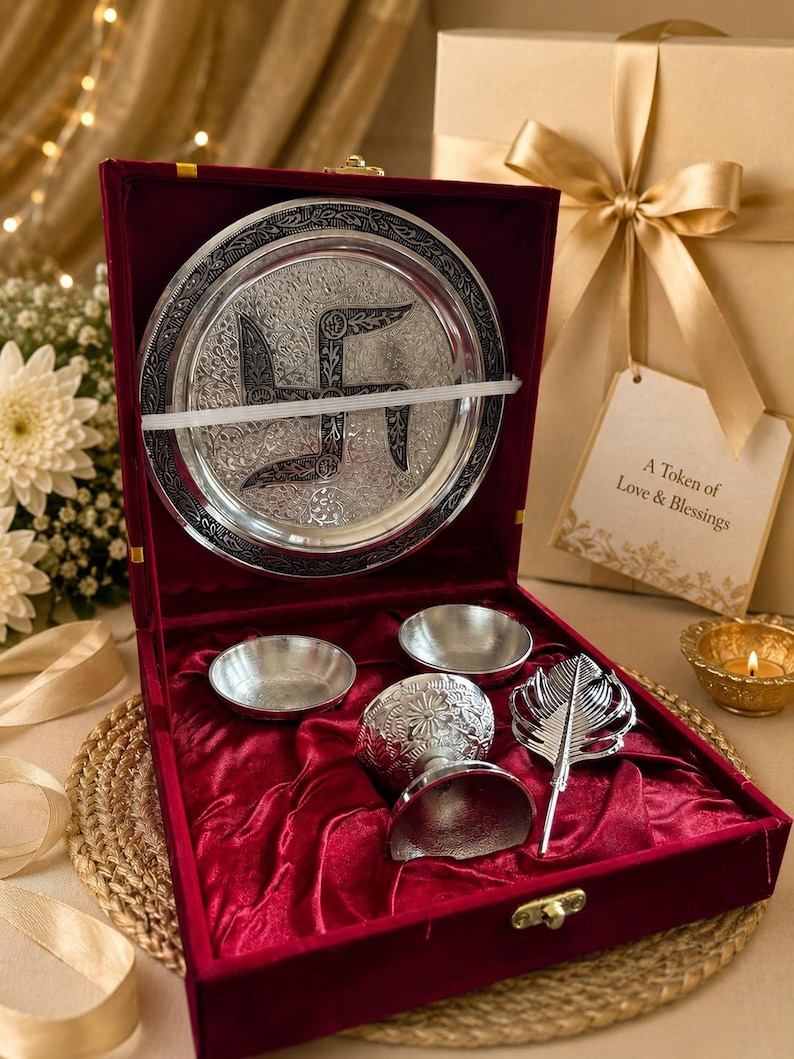 May include: A silver-toned gift set in a red velvet-lined box containing a circular plate with an engraved swastika, two small bowls, a pedestal cup, and a feather-shaped spoon. A gift tag reads A Token of Love & Blessings in black text.