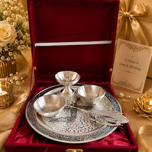 May include: A silver-toned metal puja thali set featuring a round embossed plate, two small bowls, a diya lamp, and a feather-shaped spoon, all presented in a deep red velvet-lined box with a card reading A Token of Love & Blessings.