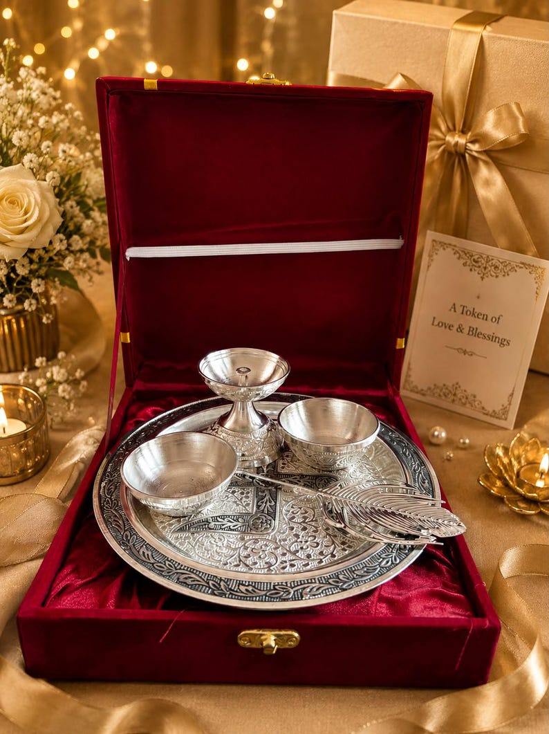 May include: A silver-toned metal puja thali set featuring a round embossed plate, two small bowls, a diya lamp, and a feather-shaped spoon, all presented in a deep red velvet-lined box with a card reading A Token of Love & Blessings.