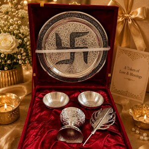 May include: A silver-toned gift set in a red velvet-lined box, featuring a circular plate with an engraved swastika symbol, two small bowls, a decorative goblet, and a feather-shaped accessory. A card reads A Token of Love & Blessings.