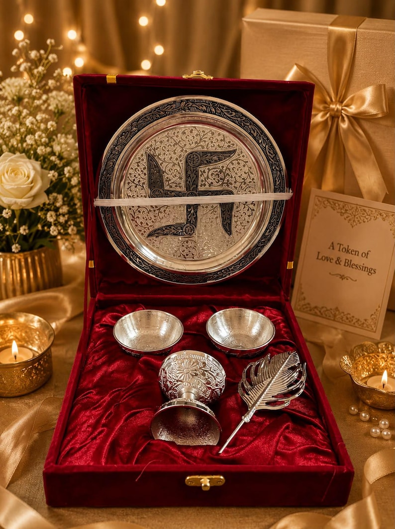 May include: A silver-toned gift set in a red velvet-lined box, featuring a circular plate with an engraved swastika symbol, two small bowls, a decorative goblet, and a feather-shaped accessory. A card reads A Token of Love & Blessings.
