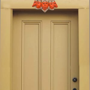May include: A decorative wall hanging featuring five orange monkey-like faces arranged in a fan shape, mounted above a light tan door. The central face is larger, flanked by two smaller faces on each side, all set against a metallic silver frame.