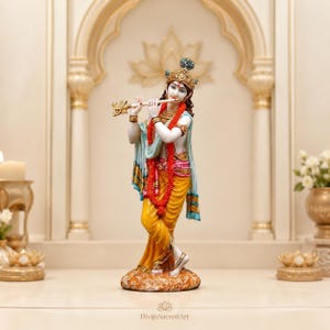May include: A standing figurine of a deity playing a golden flute, wearing a yellow dhoti, a blue shawl, and an ornate crown. The figure is adorned with red garlands and gold jewelry, standing on a textured base with the text DivijaSacredArt.