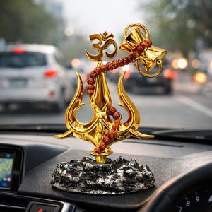 Könnte beinhalten: Goldfarbene Auto-Armaturenbrett-Ornament mit einem Dreizack, dem Om-Symbol und einem glockenförmigen Objekt. Eine Kette aus braunen Perlen hängt von der Glocke. Das Ornament steht auf einem schwarz-grau strukturierten Sockel.