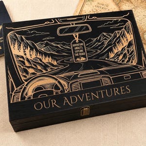 Peut inclure: Une boîte rectangulaire en bois noir avec une gravure au laser représentant l'intérieur d'une voiture face à une route de montagne. Le texte "ADVENTURE AWAITS EXPLORE THE ROAD" figure sur une étiquette suspendue et "OUR ADVENTURES" en bas.