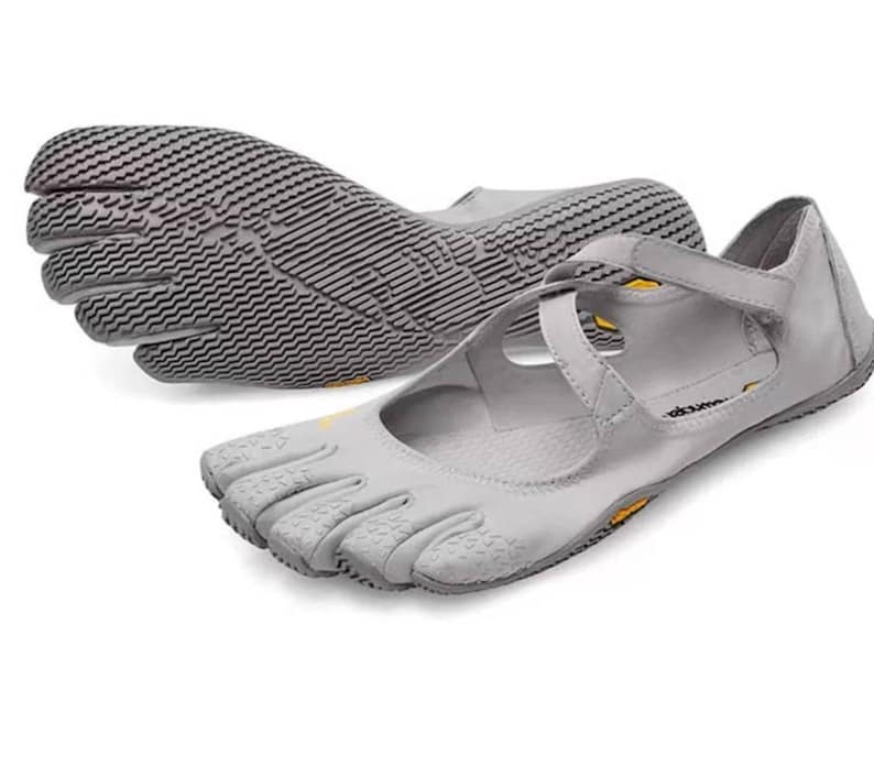 May include: A pair of light gray five-toe shoes featuring individual toe compartments, a textured rubber sole, and crisscross straps across the top. The shoes include small yellow Vibram logos on the side and the sole, with a low-profile design.