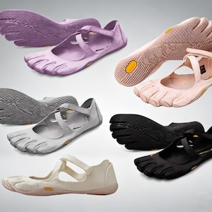 May include: A collection of five-toe shoes in purple, light pink, gray, black, and off-white. Each shoe features a flexible rubber sole with a textured grip pattern, individual toe compartments, and a cross-strap closure across the top of the foot.