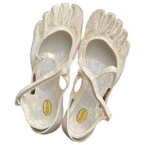 May include: A pair of off-white Vibram FiveFingers toe shoes featuring individual toe compartments, a mesh upper, and a cross-strap closure. The interior footbed displays a yellow Vibram logo, and the brand name is printed in yellow on the side.