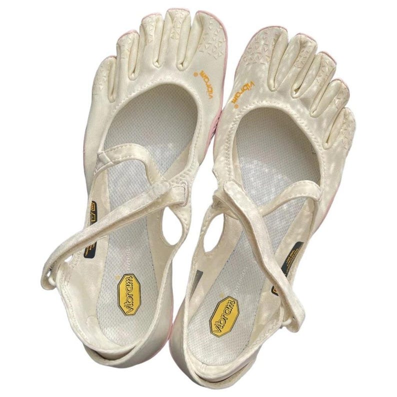 May include: A pair of off-white Vibram FiveFingers toe shoes featuring individual toe compartments, a mesh upper, and a cross-strap closure. The interior footbed displays a yellow Vibram logo, and the brand name is printed in yellow on the side.