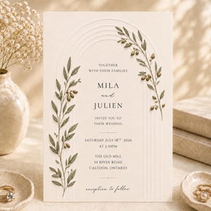 May include: A cream-coloured wedding invitation card featuring an embossed arch design and green olive branch illustrations. The text reads: Together with their families, Mila and Julien invite you to their wedding, Saturday July 18th, 2026, at 2:45PM, The Old Mill, 34 River Road, Caledon, Ontario, reception to follow.