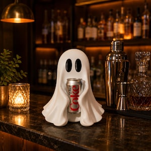 May include: A white, smooth, ghost-shaped figurine with two black oval eyes, holding a silver aluminium can of Diet Coke in its centre. The ghost has a draped, flowing appearance with rounded edges, standing upright on a dark, reflective surface.