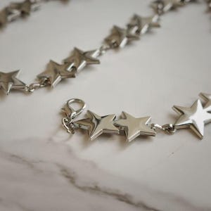 May include: A silver-toned chain necklace featuring a series of polished, five-pointed star charms linked together. The necklace includes a lobster claw clasp mechanism for secure fastening, showcasing a reflective metallic finish throughout the design.