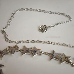 May include: A silver-toned metal chain belt featuring a series of polished star-shaped charms along the length and a single flower-shaped charm at the end of the adjustable chain, creating a reflective and metallic accessory for waist adornment.