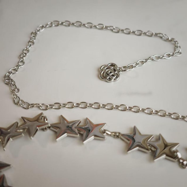 May include: A silver-toned metal chain belt featuring a series of polished star-shaped charms along the length and a single flower-shaped charm at the end of the adjustable chain, creating a reflective and metallic accessory for waist adornment.