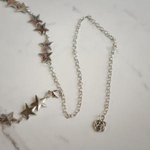 May include: A silver-toned chain necklace featuring a series of polished star-shaped charms linked together. The necklace includes a lobster claw clasp and a small, circular charm with a swirling rose-like pattern at the end of the chain.