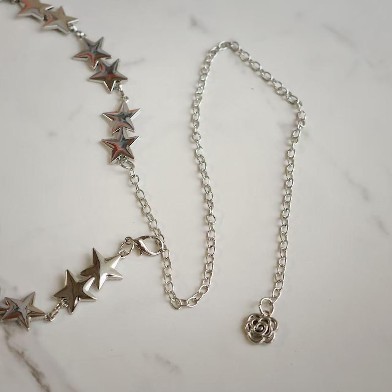 May include: A silver-toned chain necklace featuring a series of polished star-shaped charms linked together. The necklace includes a lobster claw clasp and a small, circular charm with a swirling rose-like pattern at the end of the chain.