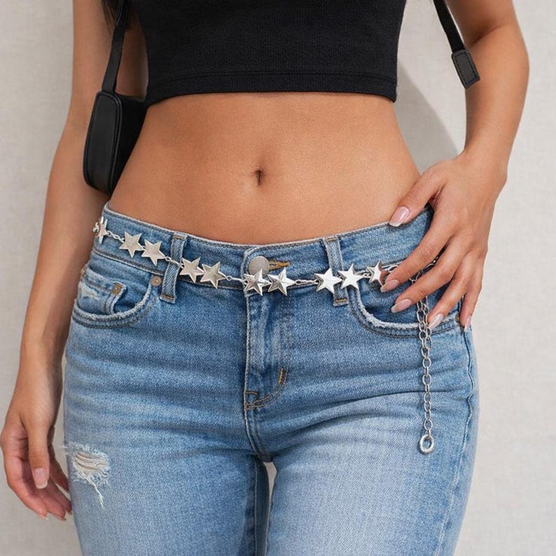 May include: A silver-toned metal chain belt featuring a series of evenly spaced star-shaped charms. The belt is worn around the waist over blue denim jeans that have a distressed detail on the left side. A dangling chain segment hangs down.