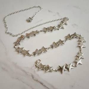 May include: A silver-toned chain belt featuring a series of connected star-shaped charms. The belt includes a thin, plain chain section with a small flower-shaped detail and a lobster claw clasp closure at the end for secure fastening.