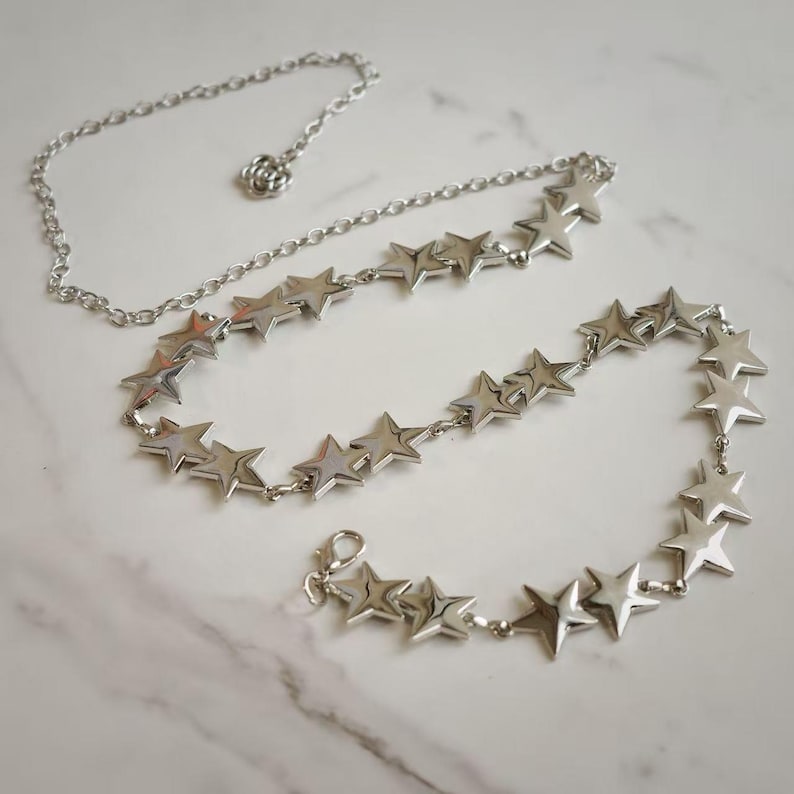 May include: A silver-toned chain belt featuring a series of connected star-shaped charms. The belt includes a thin, plain chain section with a small flower-shaped detail and a lobster claw clasp closure at the end for secure fastening.