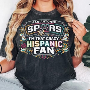 May include: Dark gray t-shirt with the text "SAN ANTONIO SPURS I'M THAT CRAZY HISPANIC FAN" in a colorful design. The design includes the Spurs logo, stars, flowers, and decorative swirls. The shirt is a crew neck.