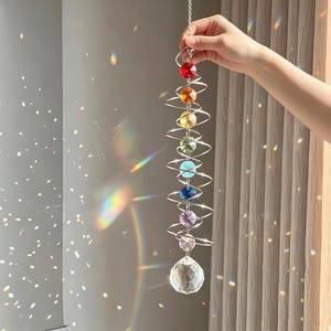 Op de afbeelding: Een decoratieve hangende kristallen zonnevanger met een regenboog van gekleurde kristallen kralen en een grote facetgeslepen kristallen bol aan de onderkant. De kralen zijn rood, oranje, geel, groen, blauw, indigo en violet. De zonnevanger wordt opgehangen door een zilveren ketting.