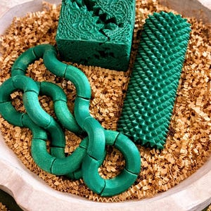May include: A wooden bowl filled with gold-colored crinkle paper holds three green fidget toys. The toys include a cube with a carved design, a textured cylinder, and a chain of connected rings. The background is a solid green.