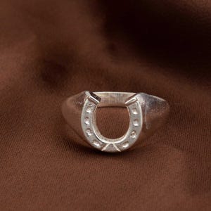 Könnte beinhalten: Ein silberfarbener Ring mit einem Hufeisen-Design. Das Hufeisen befindet sich in der Mitte des Rings und weist kleine, gleichmäßig verteilte Vertiefungen auf. Der Ring hat eine glatte, polierte Oberfläche und ein dickes Band.