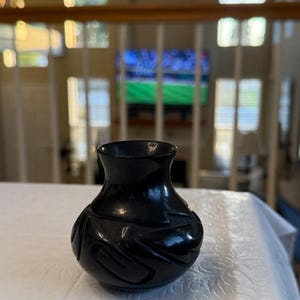May include: A small, polished black vase with a rounded body and a flared rim. The surface of the vase features carved, abstract geometric patterns that create a textured appearance across the dark, reflective material of the vessel.