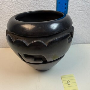 May include: A black ceramic bowl with a smooth, polished finish and carved geometric patterns around the upper section. A blue ruler is positioned behind the bowl, and a small yellow sticky note with the number 8 is placed in front.
