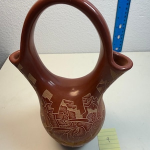 May include: A terracotta-colored ceramic wedding vase with a high arched handle and two spouts. The body features etched floral and hummingbird designs. A yellow ruler at the base shows the vessel is approximately 7 inches wide at the bottom.