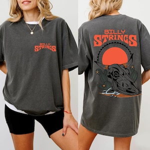 Puede incluir: Camiseta gris oscuro con el texto "BILLY STRINGS" en naranja en la parte delantera y trasera. El diseño trasero presenta una calavera de toro, cactus y un sol poniente. La camiseta tiene un corte holgado.