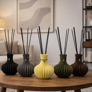 Könnte beinhalten: Fünf dekorative Schilfrohrdiffusoren in Schwarz, Grau, Gelb, Grün und Braun, jeweils mit schwarzen Schilfrohren. Die Vasen haben ein geripptes Design und stehen auf einem runden Holztisch. Der Hintergrund umfasst ein Sofa, Kunstwerke und ein Bücherregal.