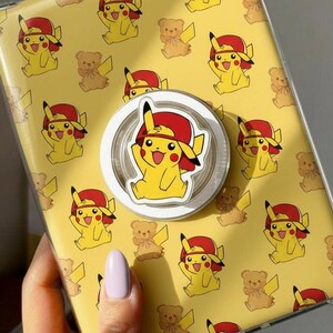 May include: Yellow tablet case featuring a repeating pattern of Pikachu in a red hat and teddy bears. A white and clear phone grip with a Pikachu design is attached to the case.