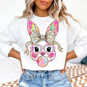 May include: White sweatshirt featuring a cartoon bunny face with heart-shaped glasses and a bubblegum bubble. The bunny has colorful ears, a polka-dot bow, and gold accents. The design is playful and whimsical.