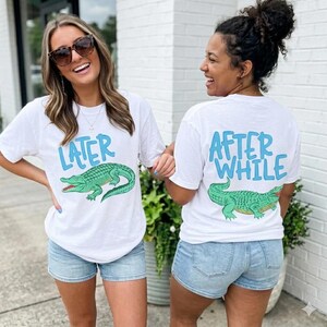 Pode incluir: T-shirts brancas com um desenho de jacaré verde. Uma camisa diz "LATER" em azul, a outra "AFTER WHILE". As camisas são usadas com calções de ganga.