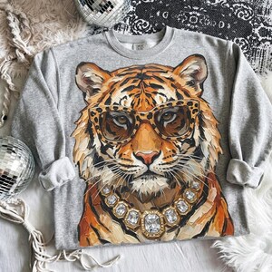 Könnte beinhalten: Ein graumeliertes Langarm-Sweatshirt mit einer großen, malerischen Illustration eines Tigers, der eine Brille mit Leopardenmuster und eine klobige Halskette mit facettierten Kristallen trägt. Der Tiger hat orangefarbenes, schwarzes und weißes Fell.