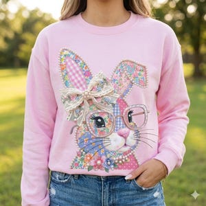 May include: A light pink sweatshirt featuring a whimsical bunny design. The bunny has floral patterned ears, a bow, and glasses. The design is embellished with pearls. The sweatshirt is a comfortable, casual style.