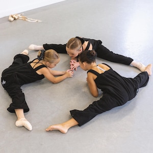 May include: Three young dancers in black jumpsuits perform the splits on a grey floor in a dance studio. Ballet shoes lie in the background. The girls are focused on something in the centre of their circle.