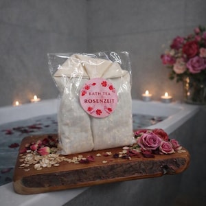 Könnte beinhalten: Ein in Klarsichtfolie verpacktes Badetee-Paket mit der Aufschrift "Bath Tea Rosenzeit" liegt auf einem Holztablett mit Rosenblättern und Haferflocken. Das Tablett befindet sich in der Nähe einer Badewanne mit Wasser und Blütenblättern, mit Kerzen im Hintergrund.