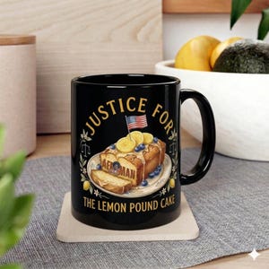 以下が含まれることがあります： 「JUSTICE FOR」と「THE LEMON POUND CAKE」の文字が入った黒いセラミックマグ。レモンスライス、ブルーベリー、小さなアメリカ国旗が描かれたケーキのデザイン。マグは明るい色のコースターの上に置かれています。