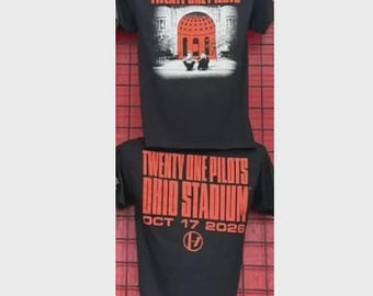 May include: A black short-sleeve t-shirt featuring the text Twenty One Pilots in orange. The top graphic shows a red domed building. The bottom graphic displays the text Twenty One Pilots Ohio Stadium Oct 17 2026 with a circular logo.
