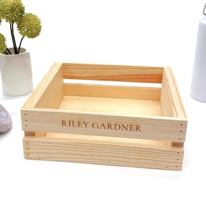 May include: A light-colored wooden crate with the name "RILEY GARDNER" engraved on the front. The crate is rectangular and has a slatted design. A white water bottle and a small vase with green decorative flowers are in the background.