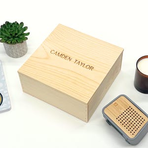 May include: A light-colored wooden box with the engraved text "CAMDEN TAYLOR" on the lid. The box is rectangular and appears to be a gift box or storage container. A small succulent plant in a gray pot is visible in the background.