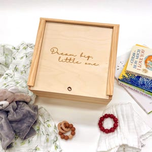 May include: A wooden box with the engraved text "Dream big, little one." is surrounded by baby items. A plush bear, a muslin blanket, books, and teething toys in shades of brown and red are visible.
