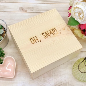 May include: A light-colored wooden box with a lid, featuring the phrase "OH, SNAP!" burned into the wood. The box is rectangular and appears to be new. The background includes flowers and decorative items.