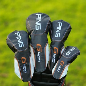 Puede incluir: Un juego de cinco fundas negras para la cabeza del palo de golf con detalles en blanco y naranja. Cada funda tiene la palabra "PING" en blanco y "G400" en naranja. Las fundas están dispuestas sobre un fondo verde.